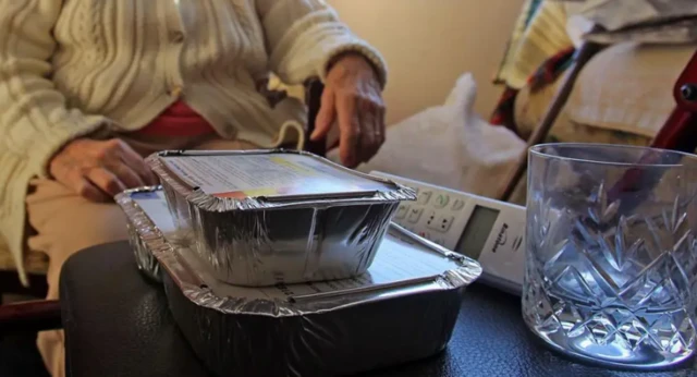 Two pre-prepared meals on a table next to a remote, with an elderly lady sat in front of them