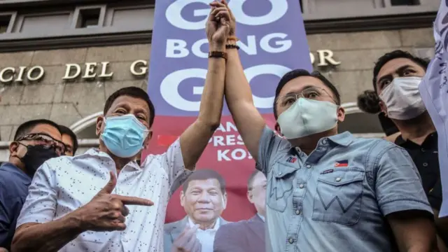 Rodrigo Duterte raises the hand of Bong Go, with both wearing face masks