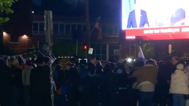 People watch a screen with an Israeli news channel on it, it is dark outside and there is Israeli flag bunting visible