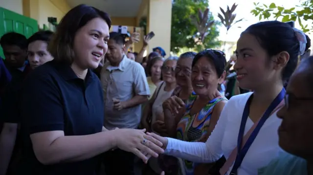 Sara Duterte in a black polo shirt shaking hands with a young woman in white. She is surrounded by other women who are smiling.