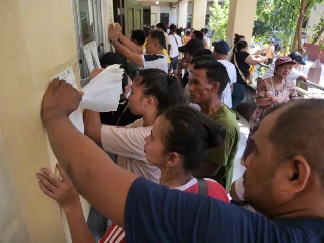 A crowd gathers in front of a yellow wall in a building. They are looking at pieces of paper pasted on the wall, and holding the attached papers with their hands