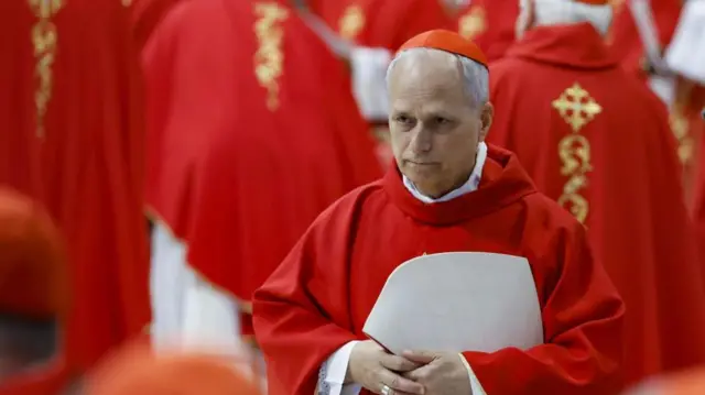 Robert prevost in red vestments as a cardinal, facing off-camera, with a crowd of cardinals behind him.