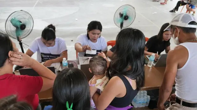 Election workers in white t-shirts sit at a long table with laptops and papers. There are standing fans blowing behind them. In front of them are people lining up to collect slips of paper from them.