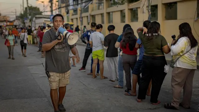 A man holding a speaker next to a queue of voters who wait to vote