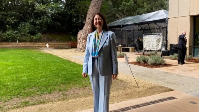 Adriana Kugler in a pale blue suit, standing outside and posing to the camera