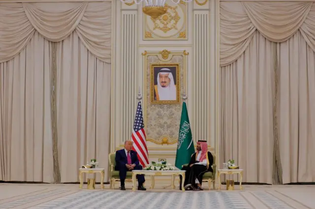Trump and MBS sitting next to each other in a large room, there is a US and Saudi flag behind them and a portrait of the Saudi king on the wall