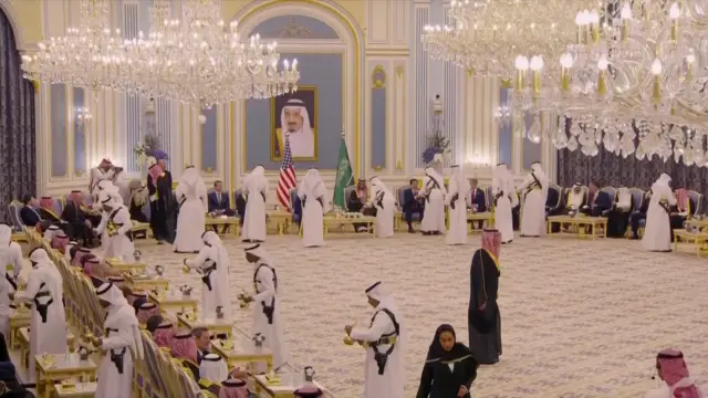 Men hand out coffee and tea in golden pots around a room, there are chandeliers and many men in suits and tradtional Saudi clothing sitting around.