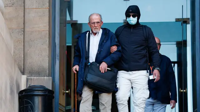 An old frail man being helped to walk down stairs by another man who wears a face mask and glasses
