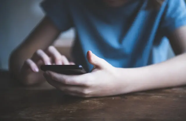 A boy in a blue t-shirt hold scrolls through a phone with both hands. His head is not visible in the picture and he is leaning on a table.