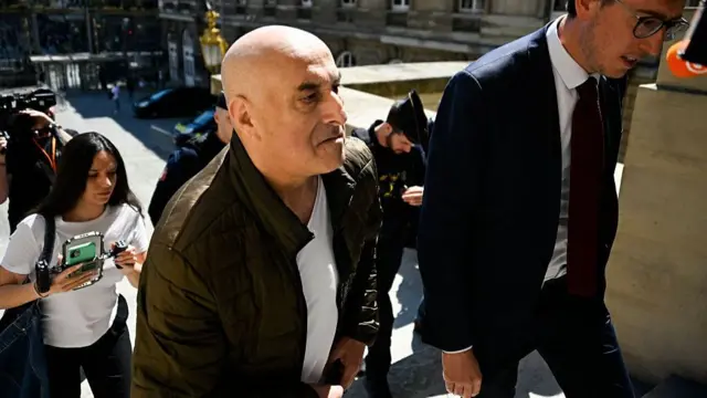 Two men walking up stairs outside with some people following them and a camera man. It's a sunny day
