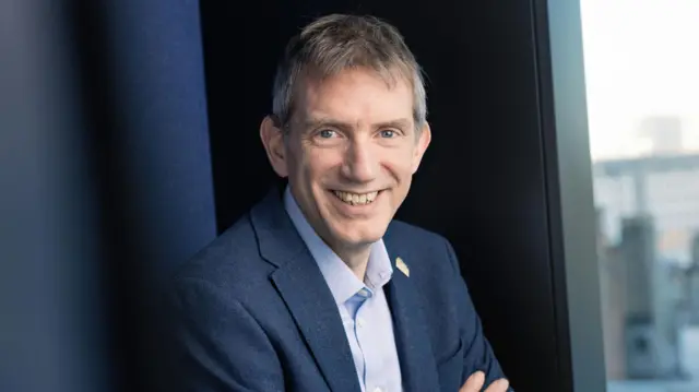 David smiles at the camera with a wide smile. He has short grey hair and wears a navy blue suit with a light blue shirt.
