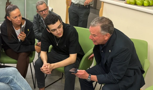 Eden Alexander, two members of his family and Steve Witkoff are sitting in chairs together. Witkoff is holding a phone which appears to be in the middle of a call.