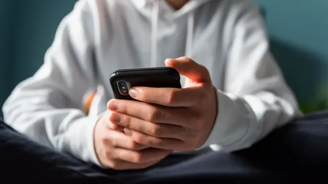 A boy in a white hoodie sits cross-legged holding a black phone.