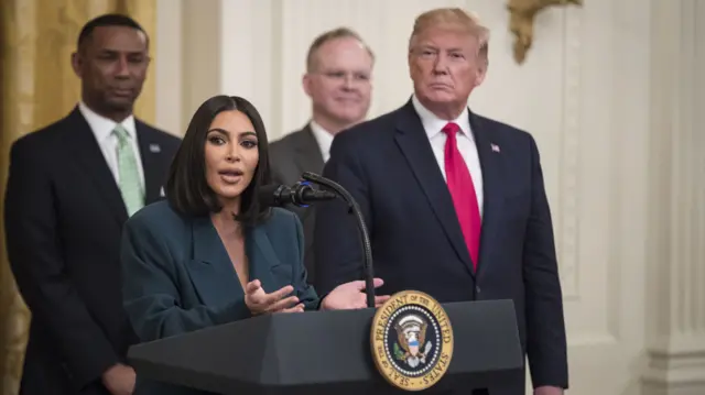 Kardashian speaking into a mic at a lectern with Trump standing behind her