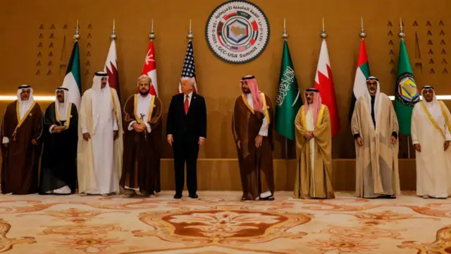 Donald Trump wears a blue suit and red tie is standing in the middle of a group of nine men, all standing in front of flags representing Gulf nations.