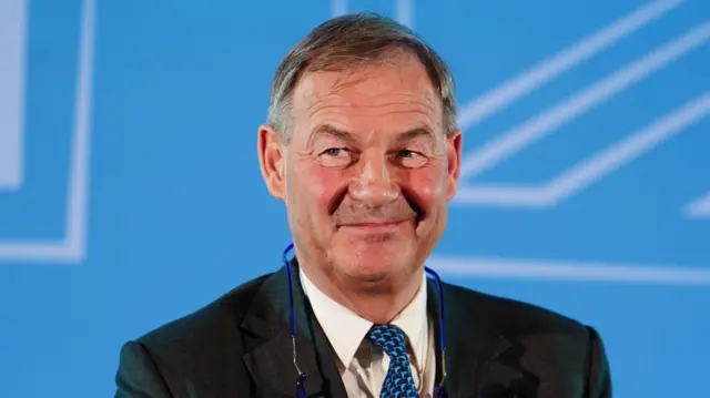 Rupert Lowe, in a suit, stands in front of a Reform UK poster at the party's conference in January