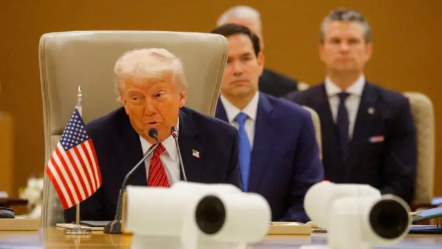 Trump sitting speaking at a table with a small US flag in front of him, and Rubio sat behind him
