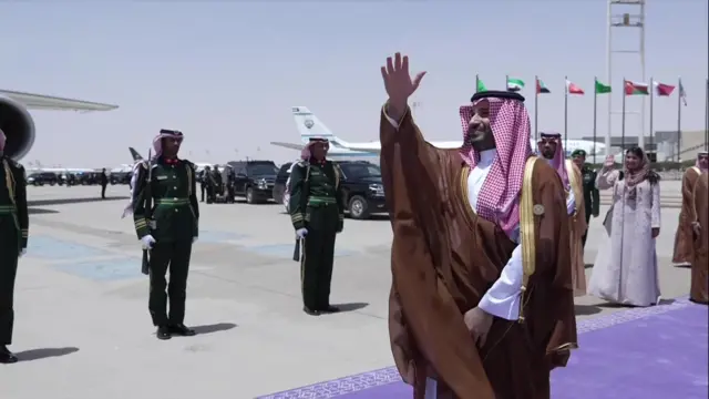 Saudi Crown Prince Mohammed bin Salman stands on a purple carpet waving at Air Force One. In the background are members of the army standing to alert.