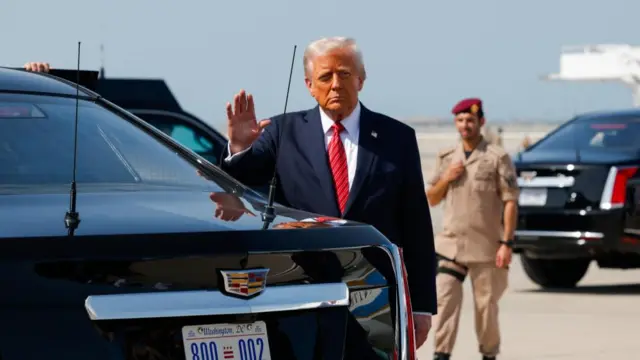 Trump waving to the camera as he stands next to a black car