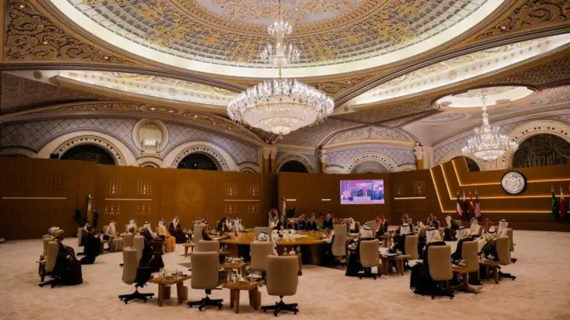 Around 60 officials sit on pale beige office chairs in a circle. All the chairs are facing a round table, where President Tump is sat. Above them are two chandeliers dangling from an ornate ceiling.