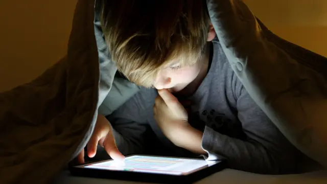 A young boy with blonde hair stares at an iPad screen. He is lying on his stomach, wearing a long-sleeved grey top, and has a grey duvet pulled over his head. His face is illuminated by the white light of the screen, and he has one thumb near his mouth.