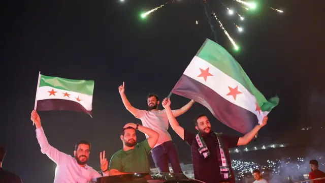 Four men stood above a car, holding two Syrian flags looking happy. A firework goes off above them filling the sky
