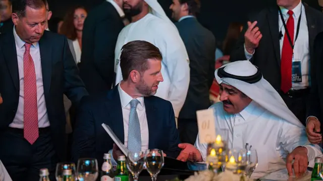 Eric Trump sat talking to a man at a table with many people behind him milling about