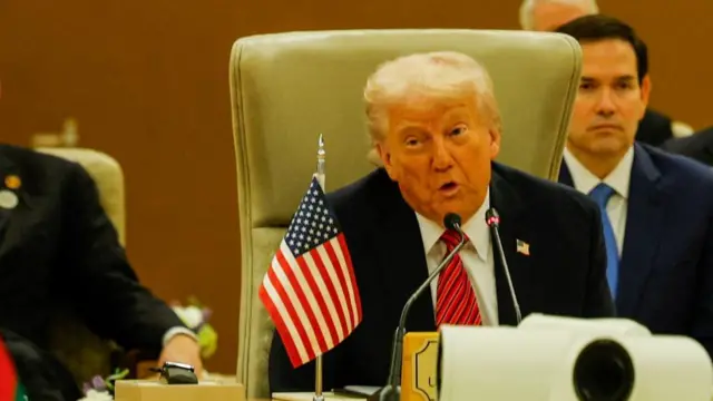 President Trump sits at a white desk speaking into a microphone. On the desk in front of him is a small US flag.