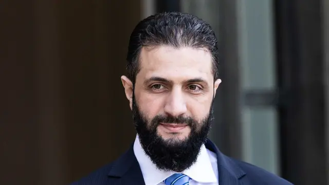 Headshot of Ahmed al-Sharaa, who smiles slightly. He has a large black beard