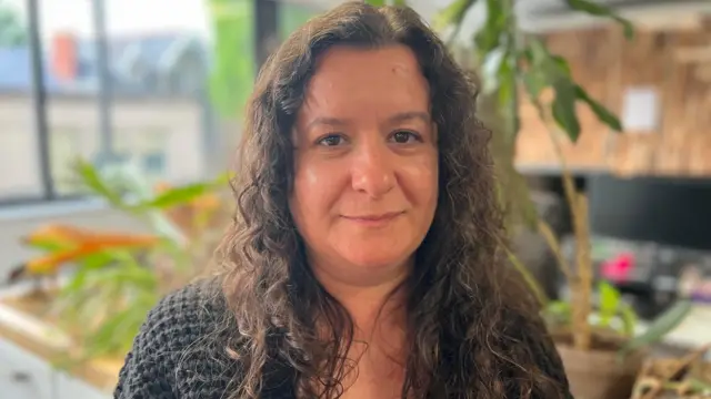 Sarah smiles with her mouth closed at the camera. She is in an office or cafe, with plants in the background. She has long dark curly hair and wears a black cardigan or jumper.