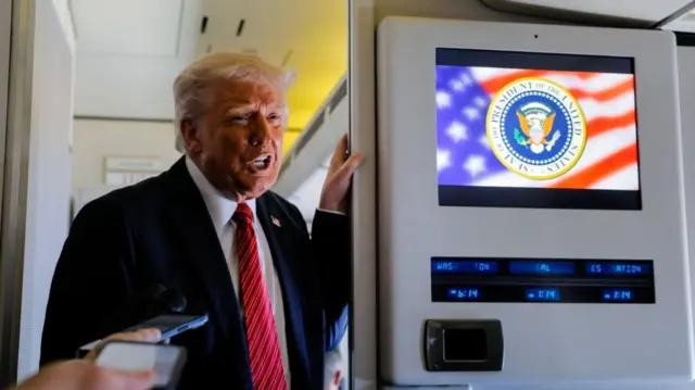 Donald Trump in black suit, white shirt and red and blue striped tie leans on the frame of a door as speaks to the press on board Air Force One
