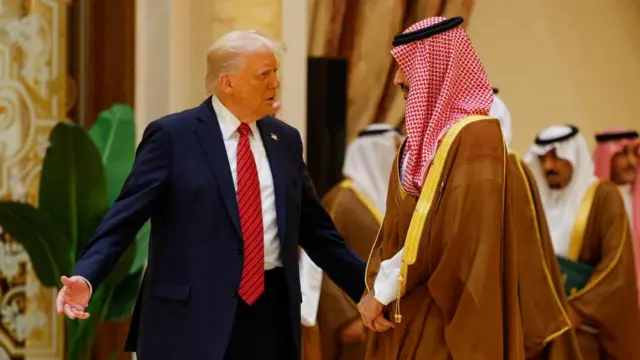 Donald Trump stands on the left of the picture facing Mohammed bin Salman whose hands are clasped in front of him. Donald Trump's arms are outstretched with his palms facing up.