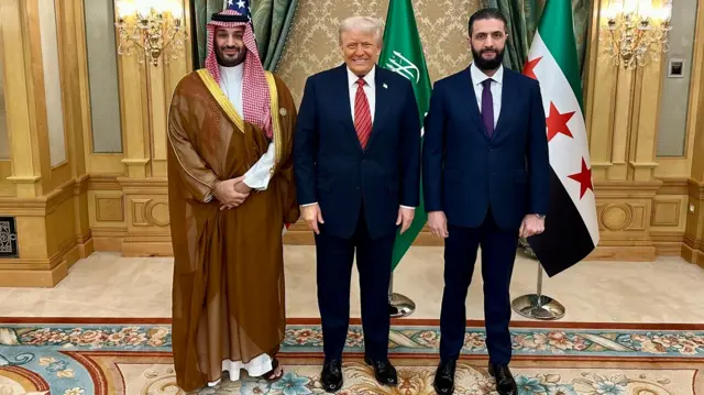 Mohammed bin Salman, Donald Trump and Ahmed al-Sharaa posing for a photo. US, Syrian and Saudi flags are behind them