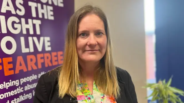 Debbie has mid-length blonde hair. She is looking straight into the camera. She is wearing a black blazer and a flowery top. She is standing in front of charity banner.