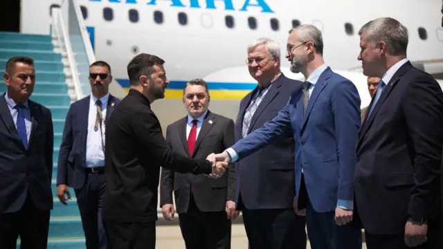President Zelensky shakes Ukrainian ambassador to Turkey Nariman Dzhelyal's hand with others standing around. A plane can be seen in the background