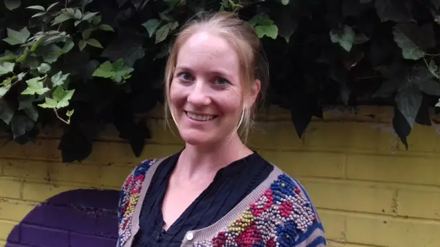 Debbie, who has blonde hair and wears a colourful cardigan over a black shirt, smiles at the camera. She is outside, in front of a yellow wall with ivy growing over it.