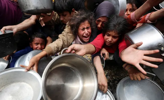 People and children hold out tin bowls and pots, crying and calling out as they wait for food