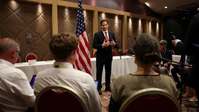 Marco Rubio stands in front of a table with a with a white table cloth covering it, and a US flag. He is speaking to people sat on chairs in a semi circle in front of him.