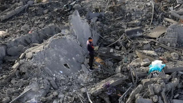 A man stands, arms folded, amid grey rubble of a destroyed house