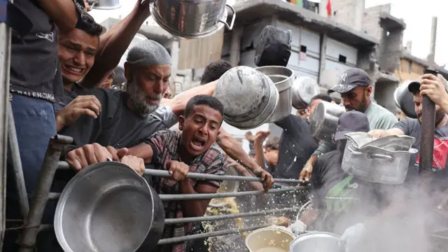 Several men holding large steel pans struggle to get food in Gaza, they are behind a metal fence.