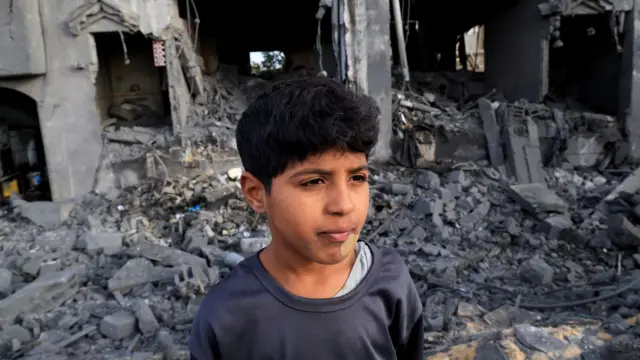 A child looks ahead, a plaster on their chin, in front of the grey rubble of a destroyed home.