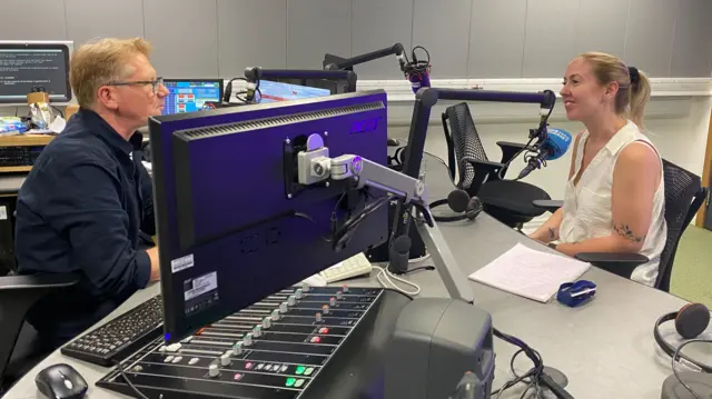 Simon, wearing a dark blue shirt, interviews Meg in a radio studio. They are both talking in to microphones, and Meg is wearing a white sleeveless shirt and has her blonde hair tied back into a ponytail.