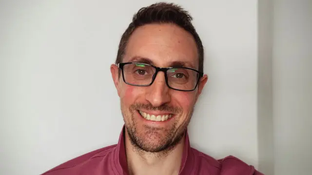 Matt Mustafic smiles at the camera. He wears a dark red shirt and square glasses, and has dark hair and stubble.