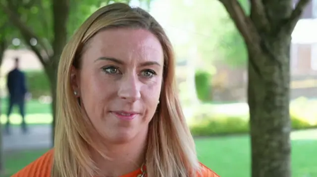 A woman wearing an orange top with blonde hair sitting infront of a blurry, glassy background