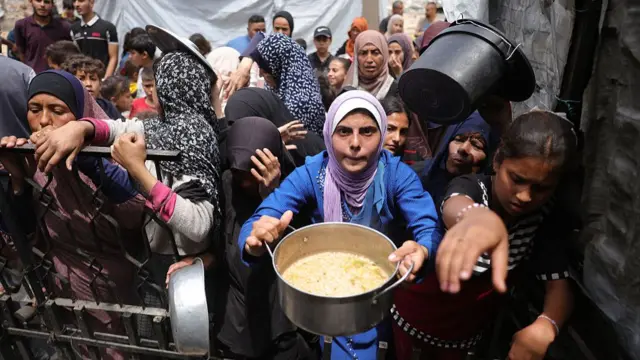 A woman dressed in blue holds out a large steel pan. Behind her a crowd has formed and another woman holds up a large black plastic bucket