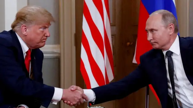 Donald Trump and Vladimir Putin sat on chairs shaking hands, US and Russian flags hanging behind them.