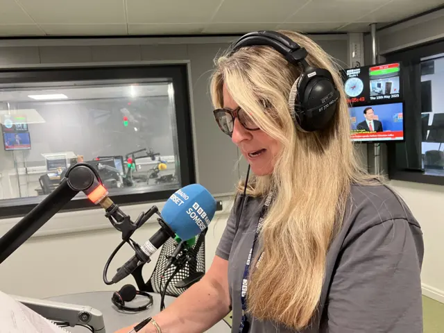 Mel sits in a radio studio, and is shown speaking into a microphone with headphones on. There is another studio visible behind her, and two television screens.