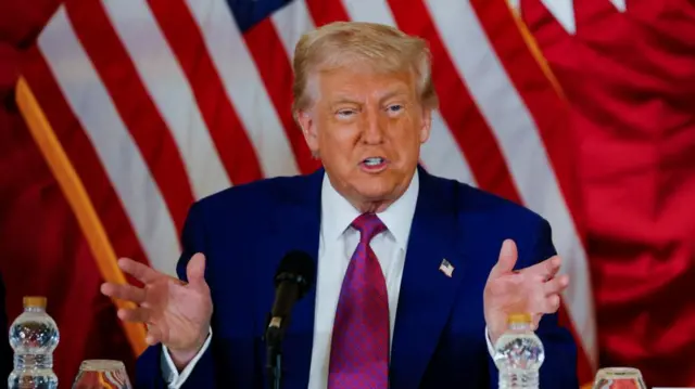Trump in a blue suit with hands raised talking into microphone.