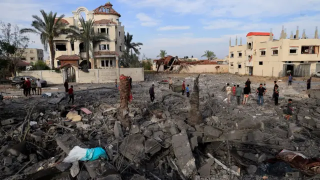 People look and walk around in a destroyed home, grey rubble on the ground beside still standing tan-coloured homes