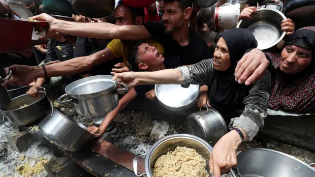 People hold out tin pots and bowls to receive food rations. A child crises, distressed.